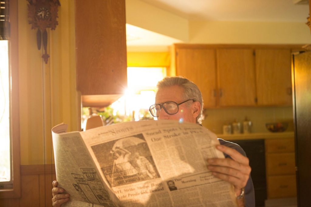 a senior man sitting in a yellow kitchen reading a newspaper while the sun streams in by lifestyle photographer Nick Reid