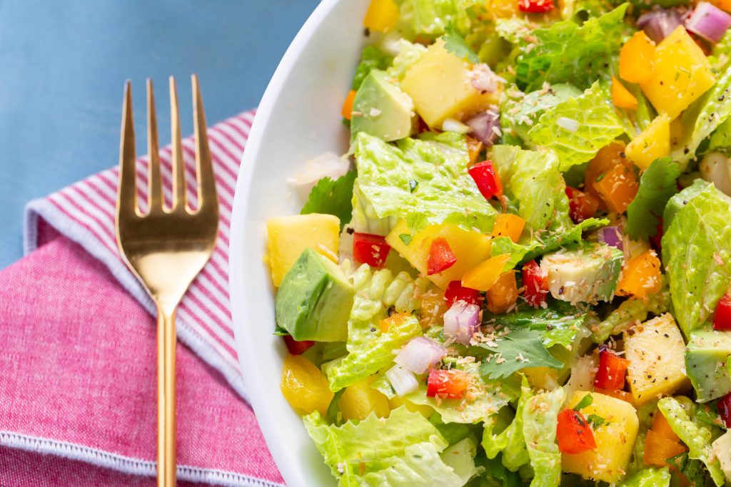 A overhead detail shot of a bright and colorful green salad with pomegranate and mango, accented by a red napkin and gold fork on a blue table.